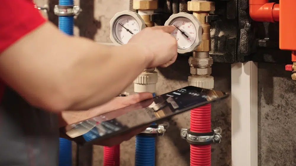 A technician in a red shirt checks pressure gauges on pipes, holding a tablet for data.