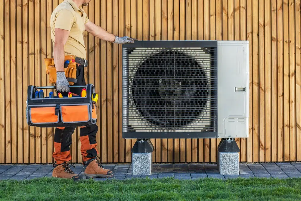 A technician stands beside an outdoor heat pump unit against a wooden wall. He holds a tool bag, wearing protective gear, conveying readiness and professionalism.