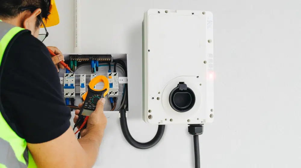 Electrician in safety gear uses a multimeter to examine wiring in an electrical panel. Nearby, a white control box is mounted on the wall.